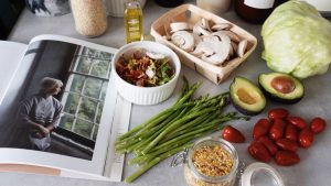 A selection of fresh vegetables and a cookbook in a kitchen setting, promoting healthy cooking.